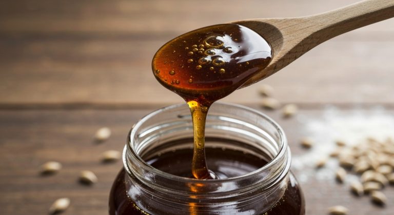 Obsahuje ječný sladový extrakt lepek? Průvodce pro celiaky 10 Hustý tmavý ječný sladový sirup stékající z dřevěné lžíce nad skleněnou nádobou na rustikální lince.